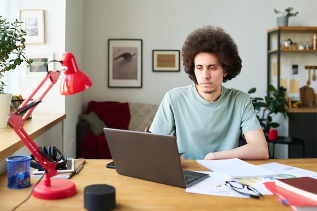 A student sitting at a desk, focused on their laptop, surrounded by textbooks and notes. A banner hangs in the background reading 